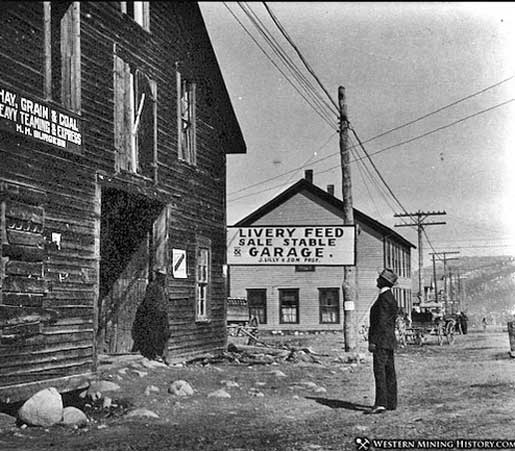 Man standing in street with horse and buggy behind him. Sign on building says Livery Feed, Sale Stable and Garage